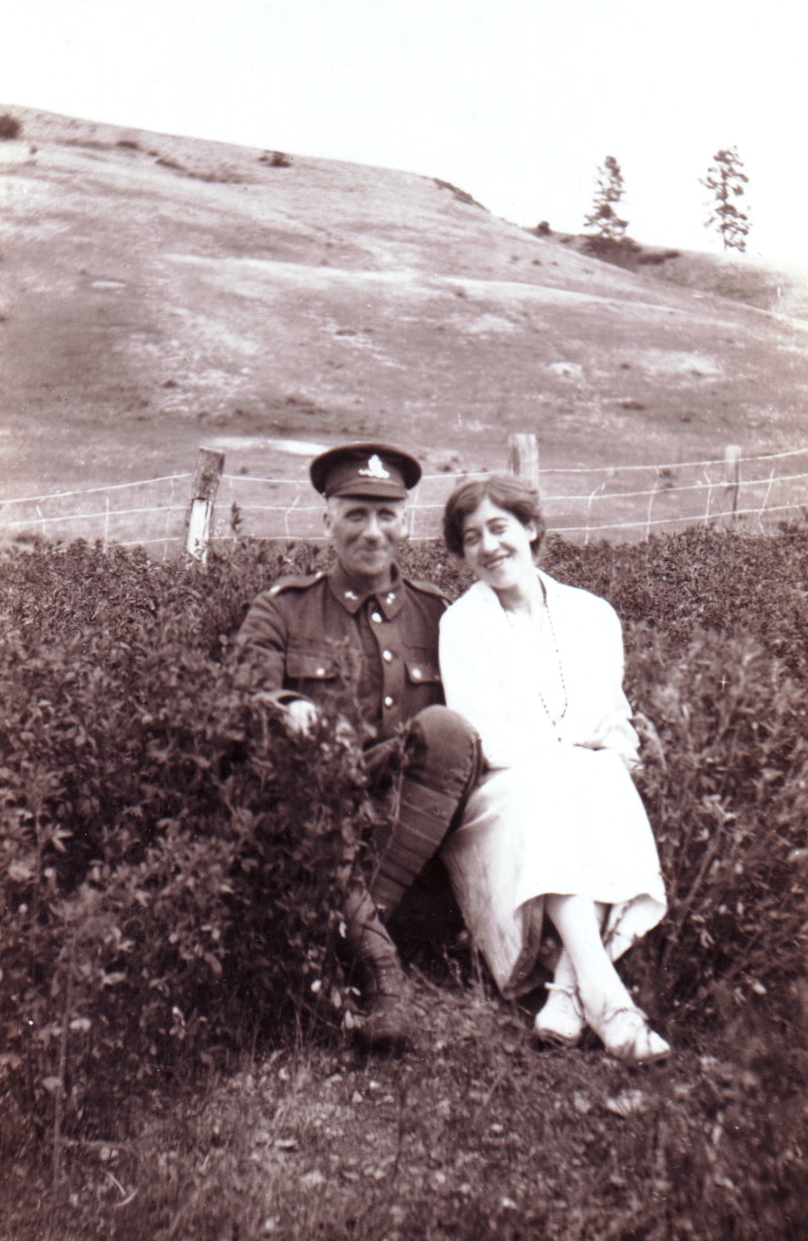 Photo en noir et blanc représentant un homme en uniforme militaire et une femme souriante assis dans un champ avec des collines en arrière-plan.