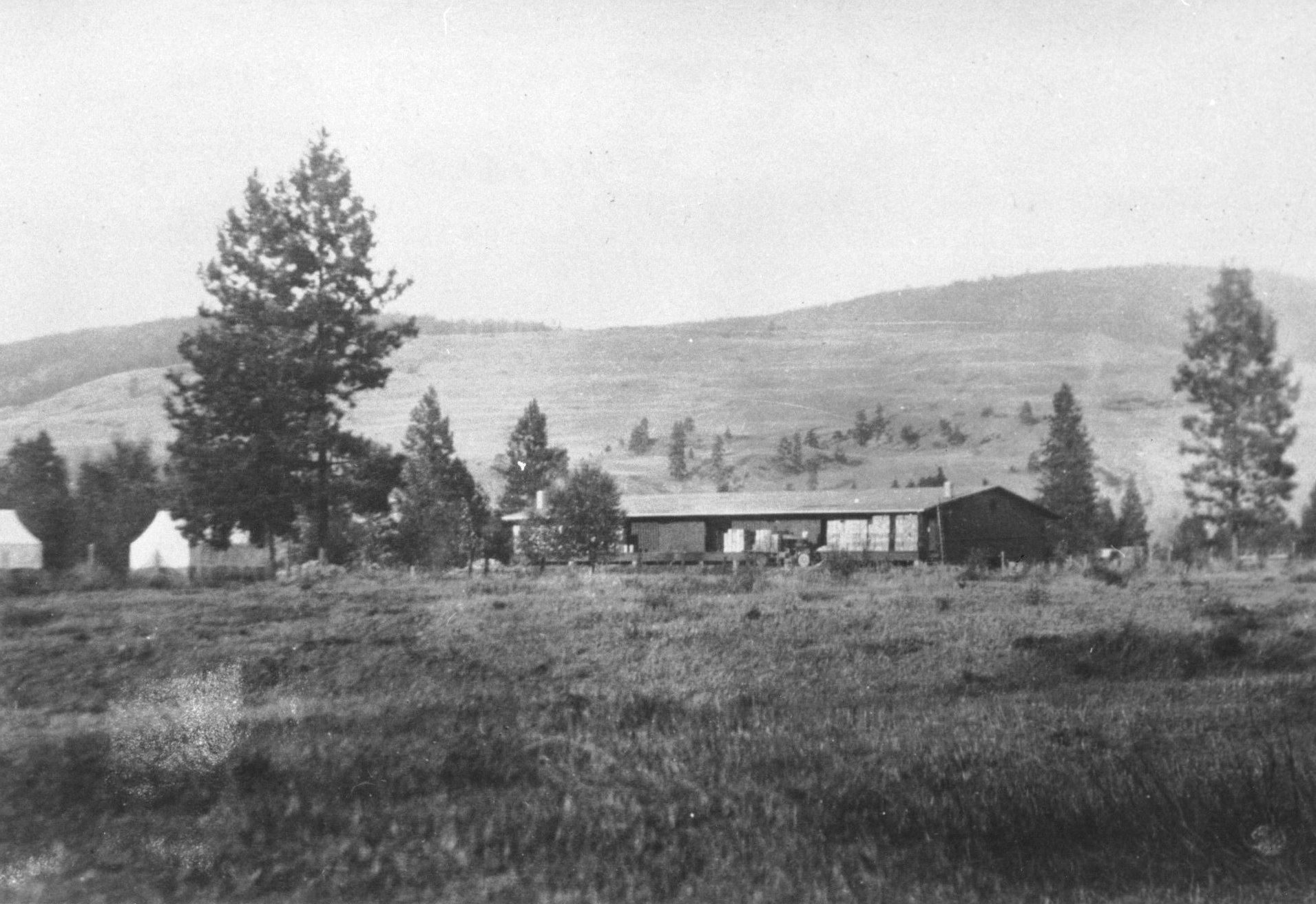 Photo en noir et blanc représentant un vieil édifice d’un étage dans un champ avec des collines en arrière-plan.