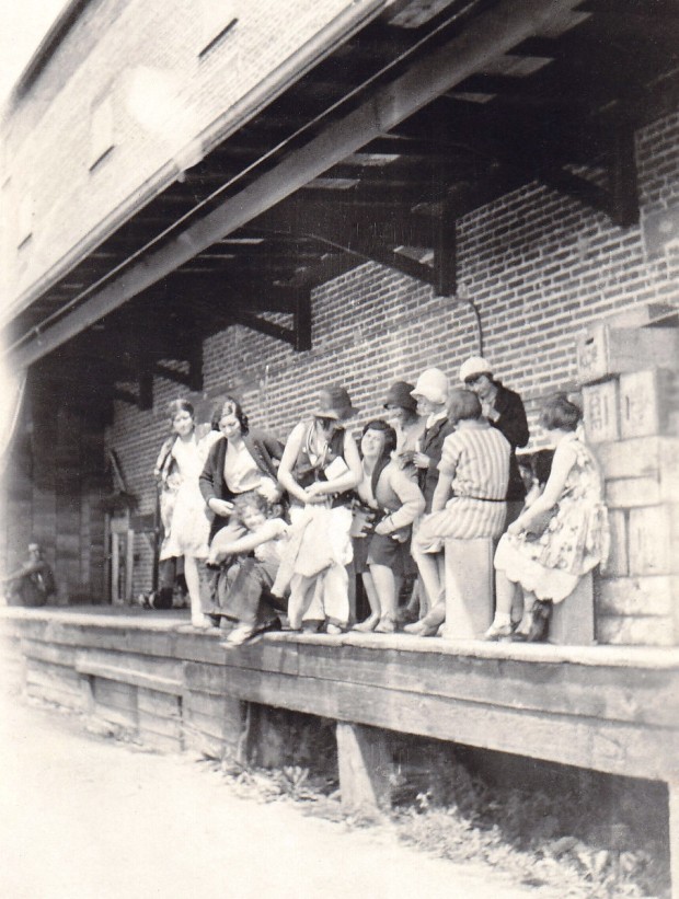 Photo spontanée en noir et blanc représentant dix femmes debout ou assises sur une terrasse de bois devant un édifice en brique.
