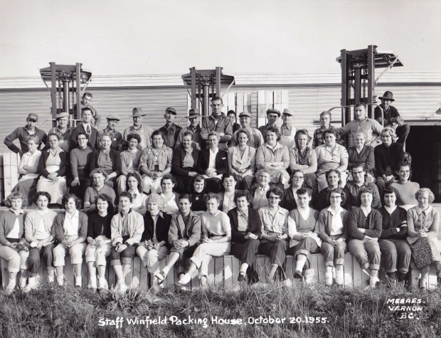 Photo extérieure en noir et blanc représentant un grand groupe de femmes et d’hommes disposé en gradins. Trois grands élévateurs à fourche et un édifice se dressent à l’arrière-plan.