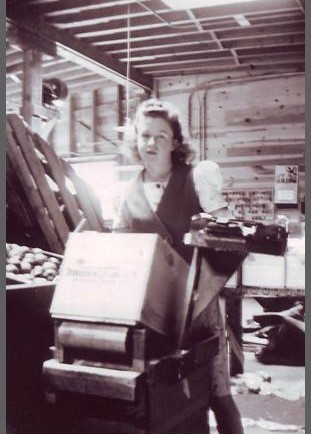 Photo en noir et blanc d’une jeune femme debout derrière une caisse de bois inclinée. Elle est à l’intérieur d’un édifice avec une benne de pommes à sa droite.