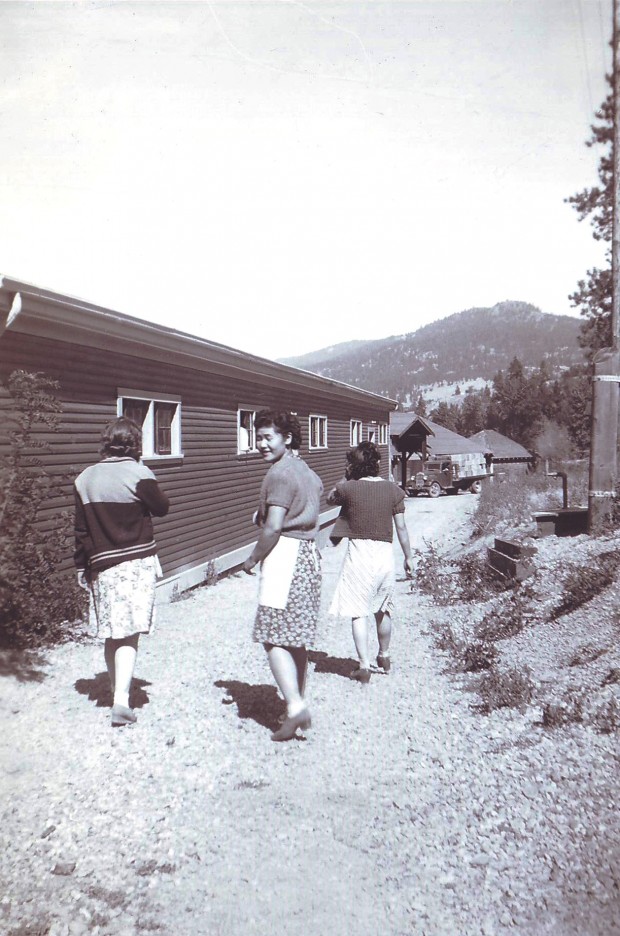 Photo en noir et blanc représentant trois femmes marchant le long d’un sentier près d’un édifice de bois d’un étage. Deux des femmes tournent le dos au photographe et la troisième se retourne pour le regarder.