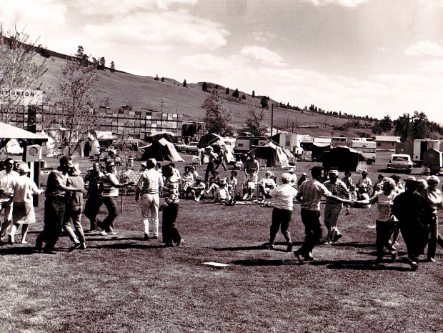 Photo en noir et blanc représentant un grand groupe d’hommes et de femmes dansant dans un champ alors que d’autres sont assis et les regardent. Derrière les danseurs, on aperçoit deux vieilles voitures, des caravanes, des tentes et des piles de caisses de pommes.