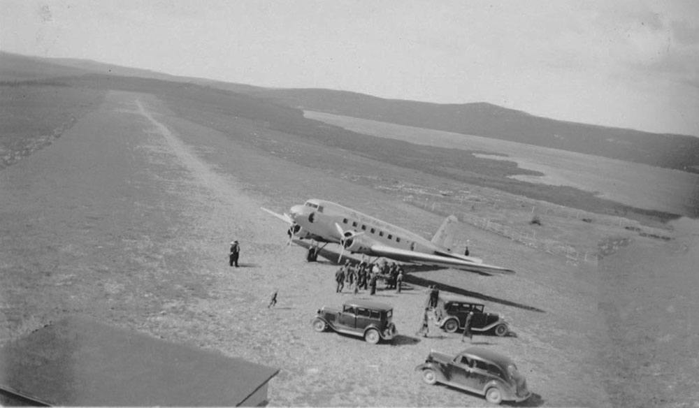 Photo en noir et blanc d’un avion Douglas DC-2 à la piste d’atterrissage, avec quelques spectateurs et trois automobiles.