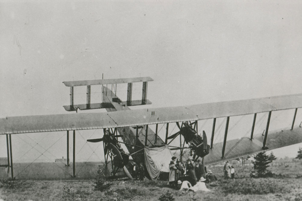 Photographie en noir et blanc du Handley Page Atlantic après son atterrissage forcé dans un champ aride, le nez vers le bas et la queue en l’air. Le nez et l’aile droite semblent endommagés. Un groupe de femmes, élégamment vêtues, se tient devant l’avion accidenté.