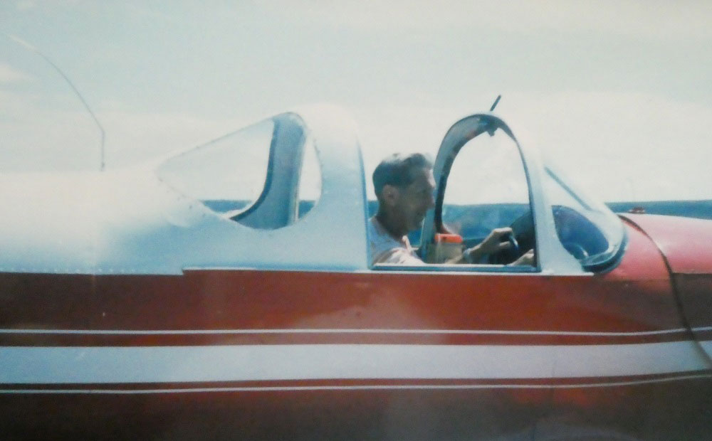 Photographie en couleurs de Claude Stevenson assis dans le poste de pilotage de son Aircoupe rouge et blanc.