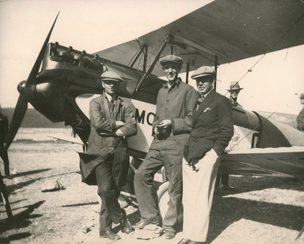 Photo en noir et blanc de trois hommes qui se tiennent devant l’avion Gypsy Moth, avec d’autres personnes en arrière-plan.