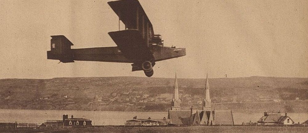 Photographie d’un journal, en noir et blanc, montrant le Handley Page Atlantic volant à environ 20 pieds au-dessus du sol lors d’un vol d’essai, avec les clochers d’église en arrière-plan, le port et la rive sud de Harbour Grace.
