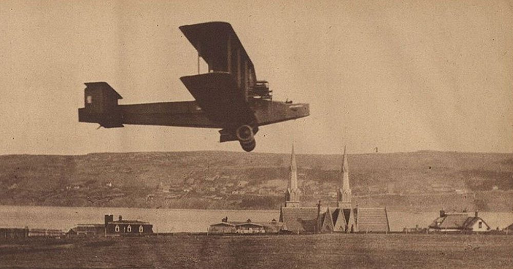 Photographie d’un journal, en noir et blanc, montrant le Handley Page Atlantic volant à environ 20 pieds au-dessus du sol lors d’un vol d’essai, avec les clochers d’église en arrière-plan, le port et la rive sud de Harbour Grace.