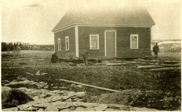 Photo en noir et blanc d’une maison en train d’être déplacée par trois hommes qui placent de gros rondins sous la maison. Il y a de nombreux rondins éparpillés sur le sol et des rochers plats.
