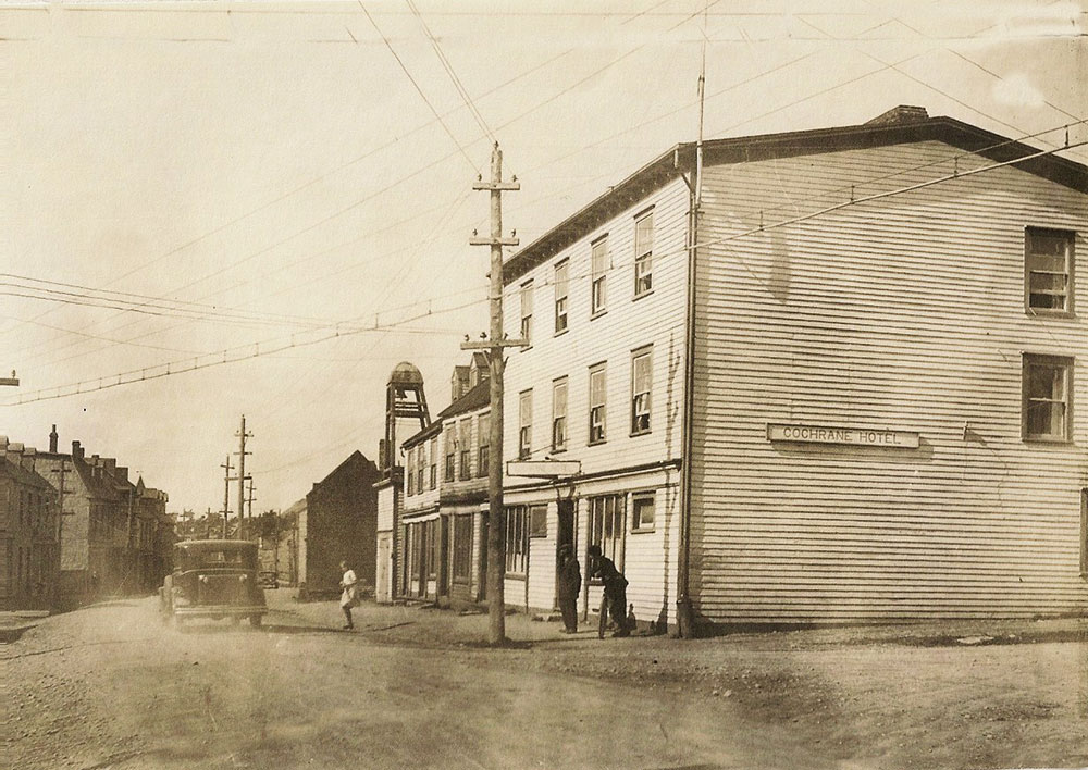 Photo en noir et blanc prise dans la rue Water à Harbour Grace, dans les années 1930, montrant un bâtiment de trois étages, l’hôtel Cochrane. Deux personnes se tiennent devant l’hôtel tandis qu’un véhicule roule sur la route.