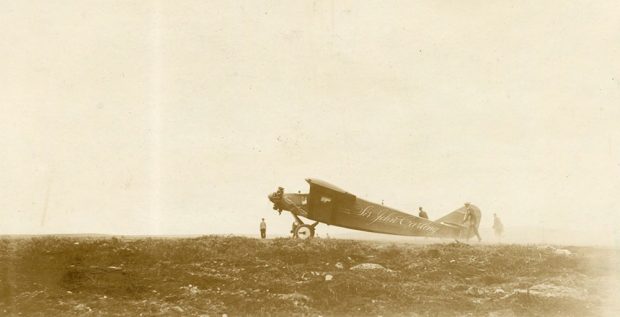 Photo en noir et blanc, vue latérale du Sir John Carling sécurisé sur la piste d’atterrissage, de loin, et entouré d’un épais brouillard. Quatre personnes se tiennent autour de l’avion.