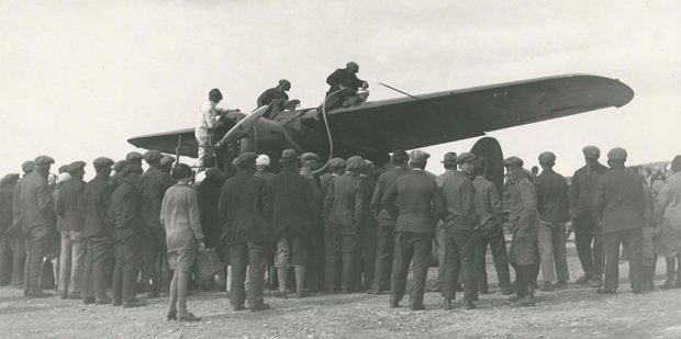 Photo en noir et blanc de trois personnes se trouvant sur les ailes d’un avion, lors du ravitaillement. Une foule nombreuse se tient autour de l’avion, dos à l’appareil-photo.