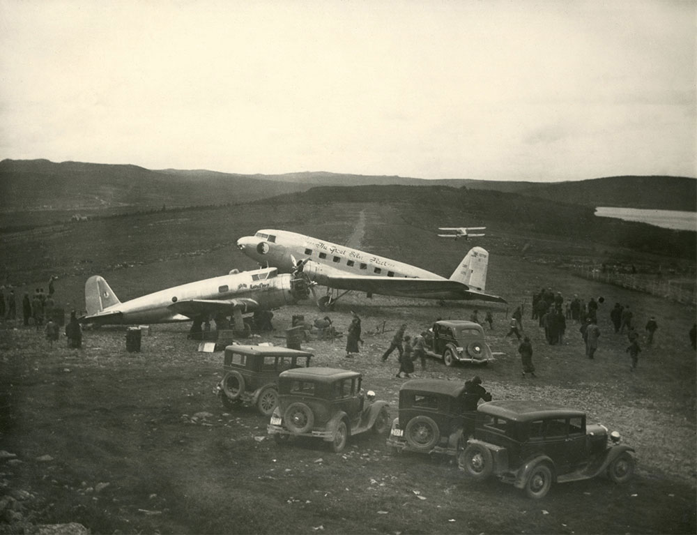 Photographie en noir et blanc montrant trois avions stationnés sur la piste d’atterrissage. Cinq automobiles sont garées tout près et une petite foule est rassemblée.