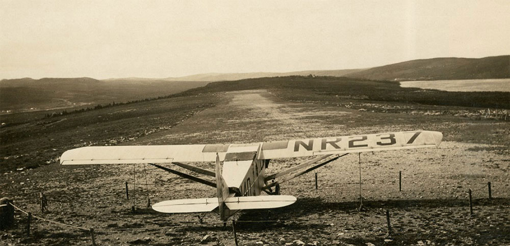 Photographie en noir et blanc de l’avion Columbia sécurisé au pied de la piste d’atterrissage en terre battue. Le numéro de repère NR237 est visible sur l’aile droite de l’avion.