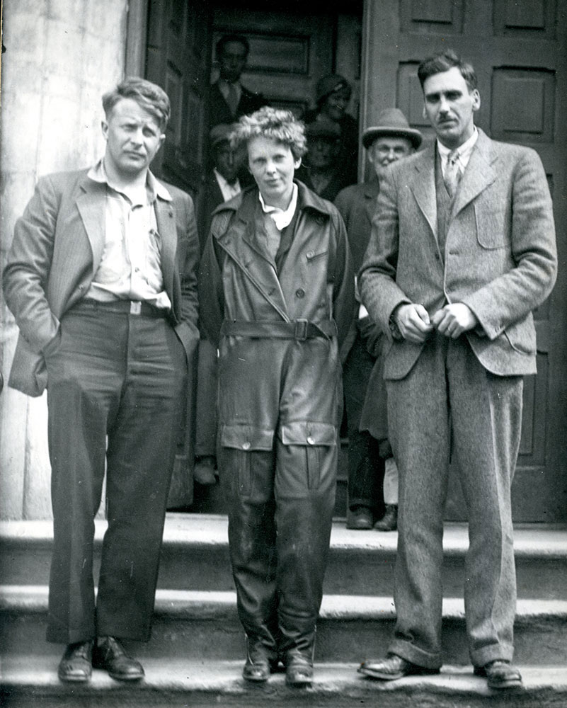 Photographie en noir et blanc d’Amelia Earhart, dans sa combinaison de vol, photographiée sur les marches extérieures du bureau de poste de Harbour Grace, un homme de chaque côté, avec d’autres personnes à l’arrière-plan.