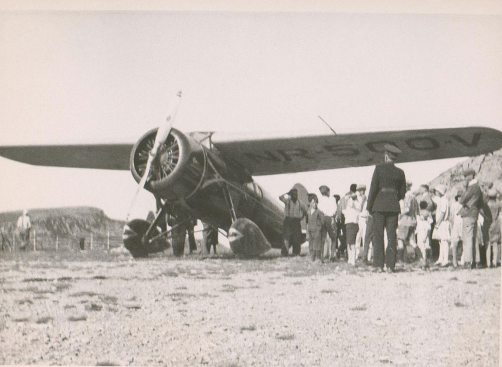 Photographie en noir et blanc de l’avion City of New York sécurisé sur la piste d’atterrissage, entouré d’une foule, notamment un homme en uniforme, possiblement un policier. Le numéro de repère NR-500-V est visible sous l’aile droite de l’avion.