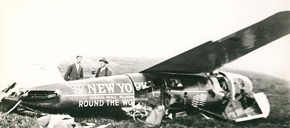 Photographie en noir et blanc de deux hommes debout derrière l’avion City of New York, après l’écrasement sur la piste d’atterrissage de Harbour Grace. L’avion est très endommagé sur le côté droit de la photo.
