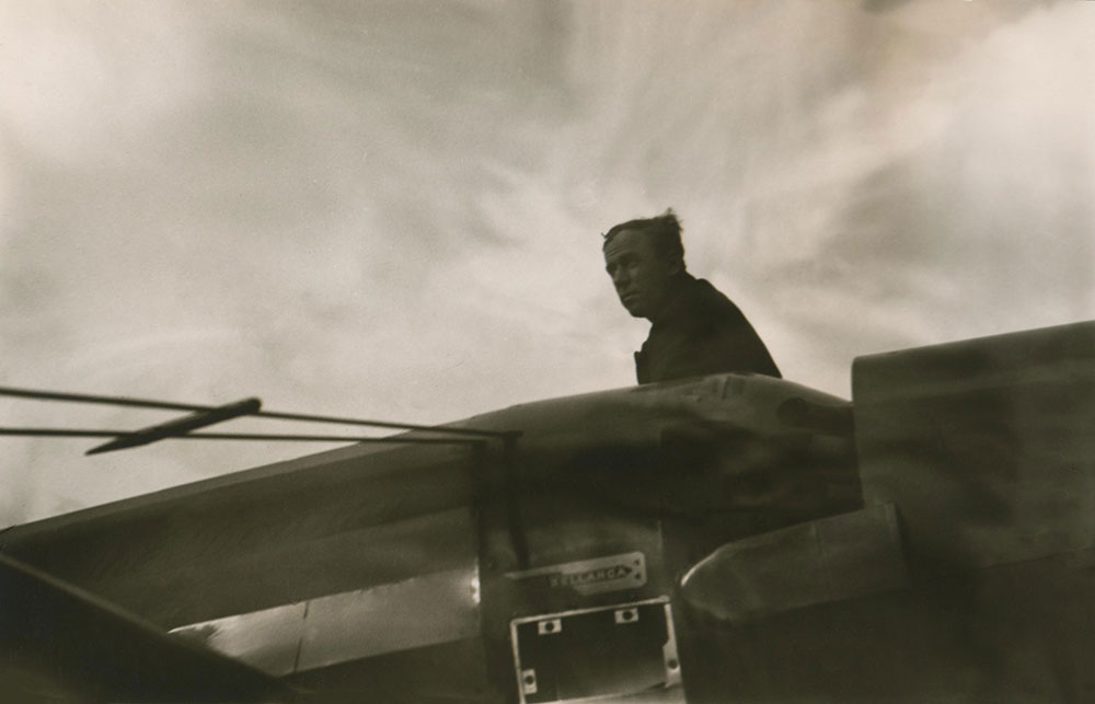 Photo en noir et blanc du pilote James Mollison assis sur le dessus de son avion, le Dorothy sur la piste d’atterrissage.