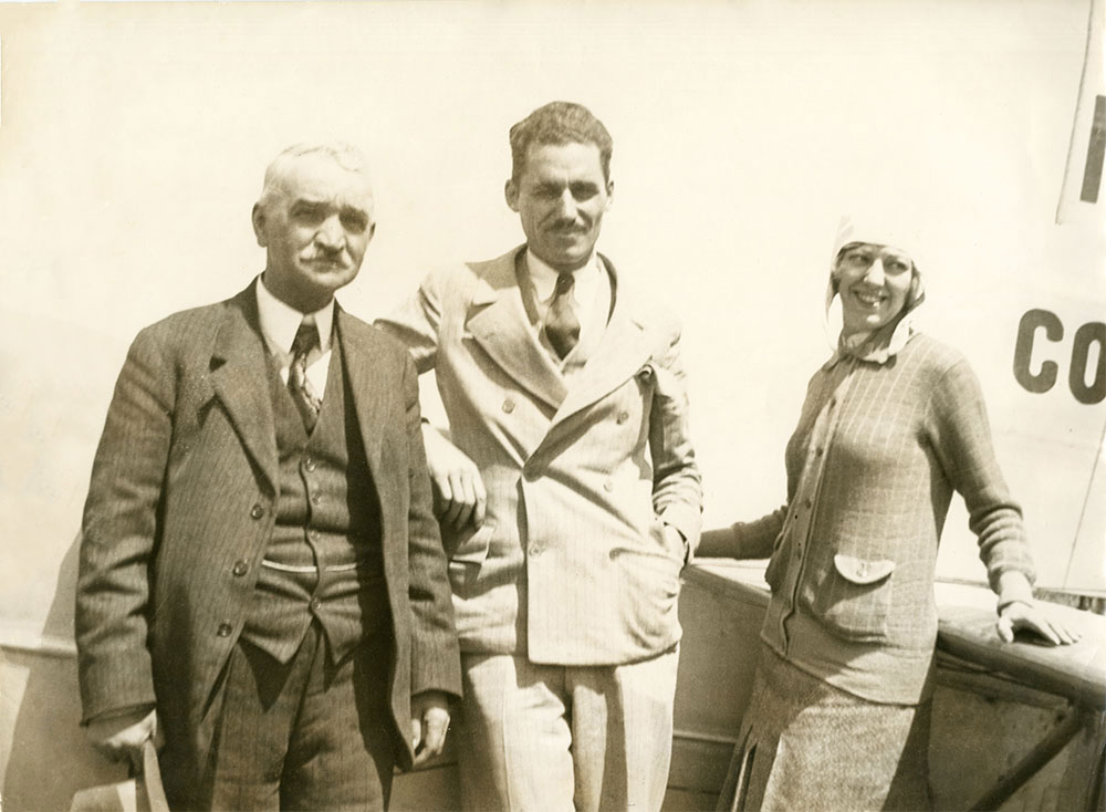 Photographie en noir et blanc de trois personnes se tenant devant l’avion Columbia. Mabel Boll, à l’extrême droite, s’appuie sur l’aile de l’avion. Les deux hommes portent des complets. Mme Boll porte un casque d’aviateur blanc, un chandail et une jupe.