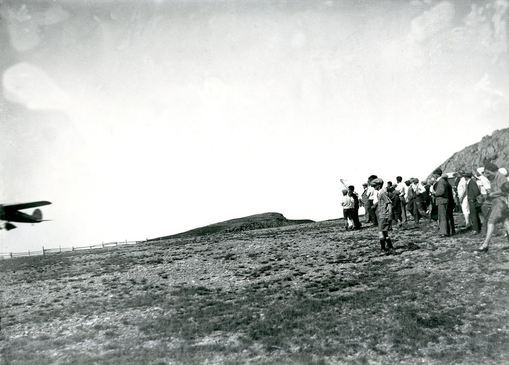 Photo en noir et blanc du Lockheed Vega d’Amelia Earhart au départ de la piste d’atterrissage de Harbour Grace. La moitié de l’avion est visible au moment du décollage, à environ 30 pieds dans les airs. Une foule est rassemblée à droite de la photo pour assister au décollage.