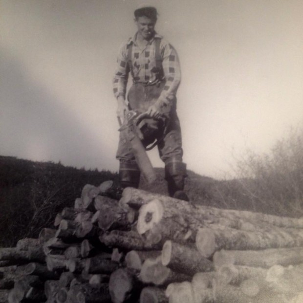 Wilson King  debout sur une pile de bois, une scie à chaine à la main 