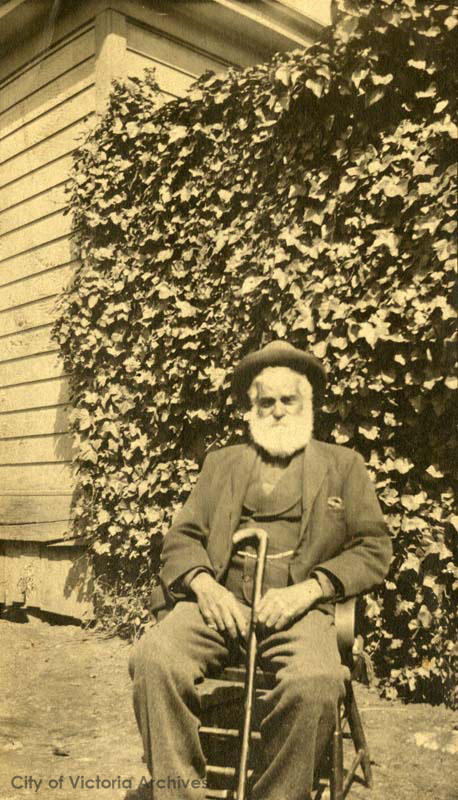 photo aux tonalités sépia, homme à un âge avancé, assis, une canne appuyée sur sa jambe. En arrière-plan, une maison et une haie de lierre