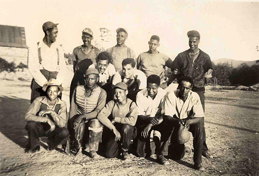 photo aux teintes sépia. Douze adolescents et jeunes adultes rassemblés en trois rangées irrégulières, debout, accroupis ou agenouillés. Habillés de façon décontractée et variée, souriants, portant chapeaux et casquettes