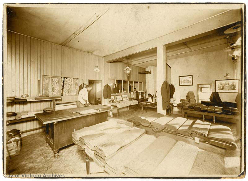 photo en noir et blanc montrant, à l’intérieur d’un bâtiment, plusieurs tables exhibant de nombreux rouleaux de tissu. Un homme se tient debout derrière un comptoir