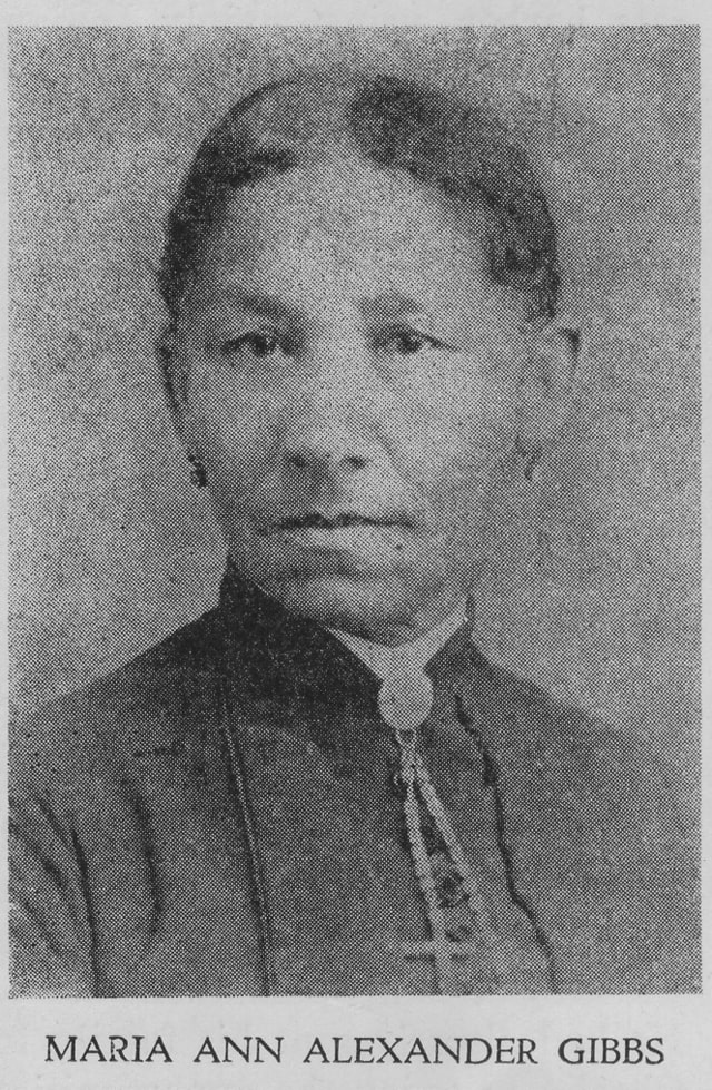 portrait professionnel en noir et blanc, vue de face d’une femme, cheveux tirés vers l’arrière et portant une robe de couleur sourde avec col montant, broche et collier