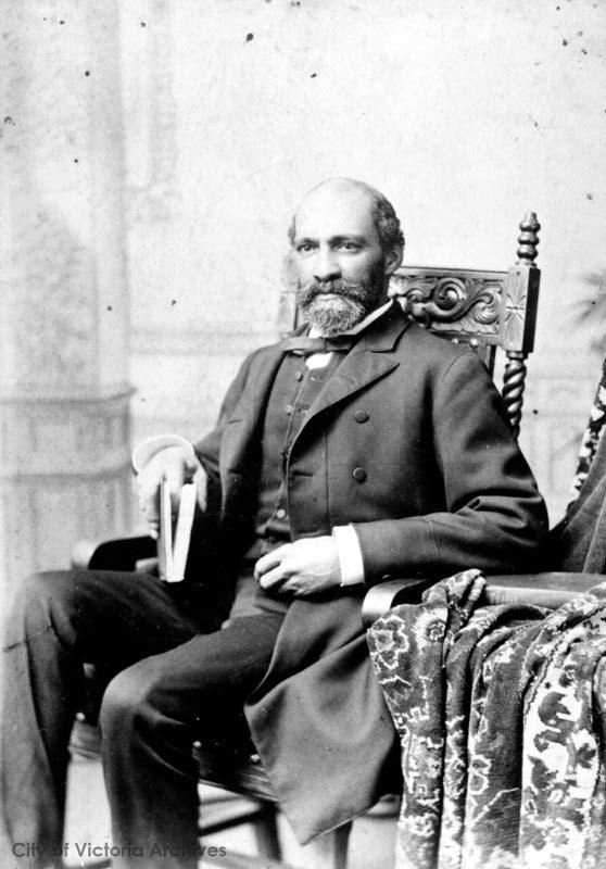 portrait studio professionnel en noir et blanc d’un homme d’âge moyen, assis, cheveux foncés et barbe taillée, portant un costume formel