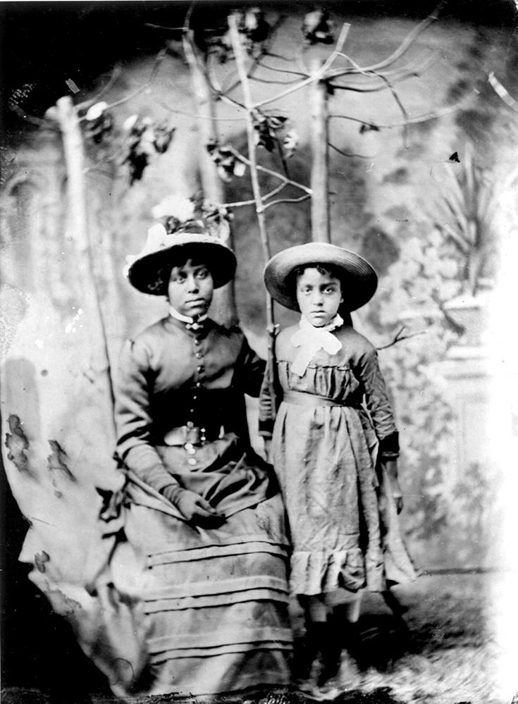 portrait professionnel en noir et blanc des deux sœurs en robe coloniale et portant un chapeau, celle de 23 ans étant assis à gauche et celle de 11 ans étant debout à côté, des arbustes feuillus occupant l’arrière-plan