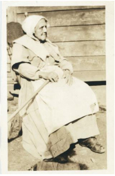 photo sépia. Une vieille femme porte un bonnet et une longue tenue de travail et un tablier, dans une berceuse sur la véranda, tenant de la main gauche un râteau sur ses genoux