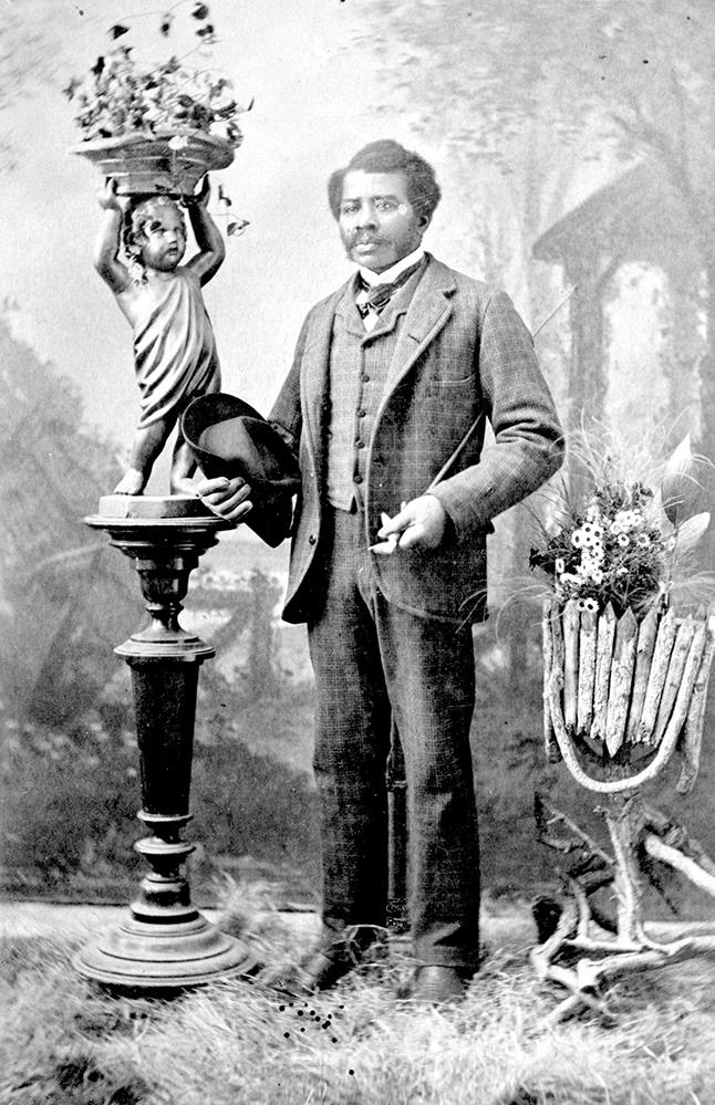 portrait studio professionnel en noir et blanc d’un homme arborant des favoris avec costume et veste, debout, tenant un chapeau et une canne; arbres et jardin classique en arrière-plan