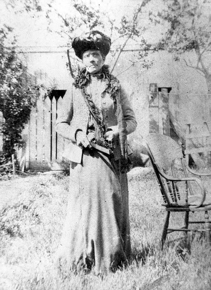 photo professionnelle en noir et blanc d’une femme se tenant majestueusement dans un jardin, portant un chapeau Victorian étagé orné de dentelle et de fleurs, une longue robe évasée de couleur claire, avec une veste de costume longueur hanches et un revers à volants