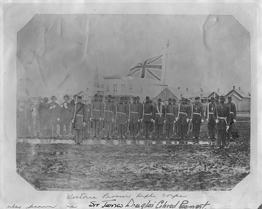 Une vingtaine de membres du « Pioneer Rifles Corp » de Victoria se tiennent debout en rangs avec le drapeau britannique en arrière-plan