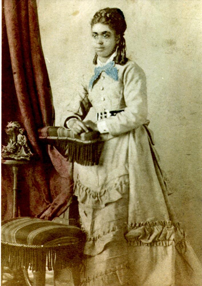 photo colorée à la main, vue de face d’une jeune femme adulte debout à côté d’une chaise, un léger sourire aux lèvres; elle porte une longue robe de couleur claire avec une large ceinture, des volants sur la jupe, et a les cheveux frisés