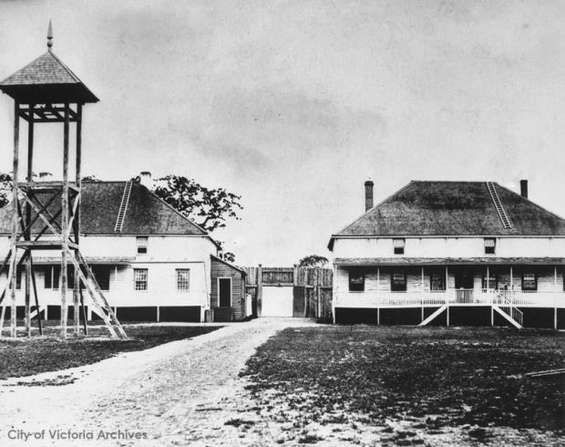 photo en noir et blanc, une tour d’observation se dresse devant deux grands bâtiments. Une porte située entre les deux s’ouvre sur un chemin passant entre les bâtiments