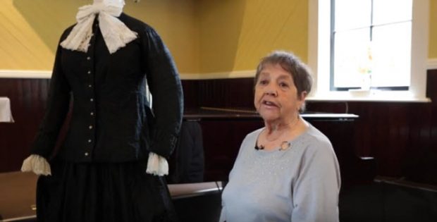 femme âgée assise sur un banc de piano dans une église, tournée vers la caméra; mannequin à sa droite arborant une longue robe noire de style colonial