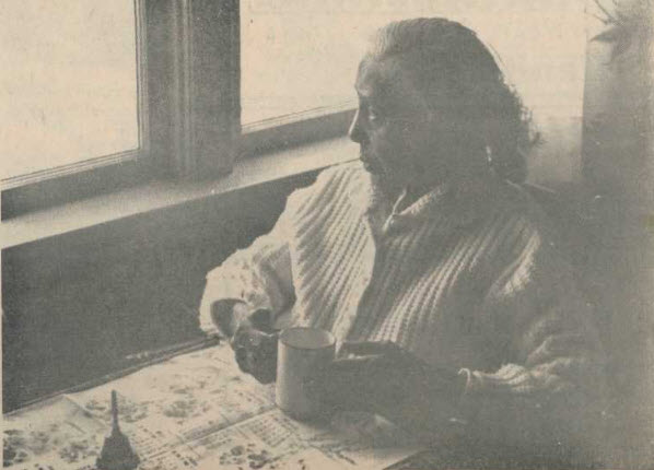 photo sépia. Une vieille femme est assise à table devant une tasse de thé et un journal ouvert, regardant par la fenêtre