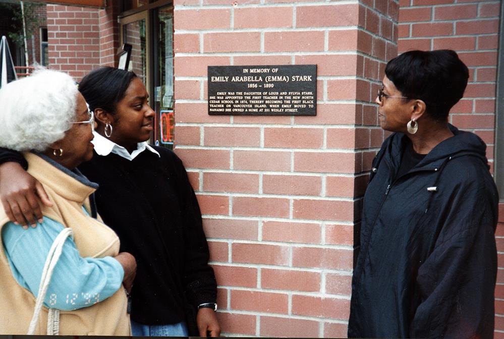 trois femmes d’âges différents fixant une plaque apposée sur un mur de briques