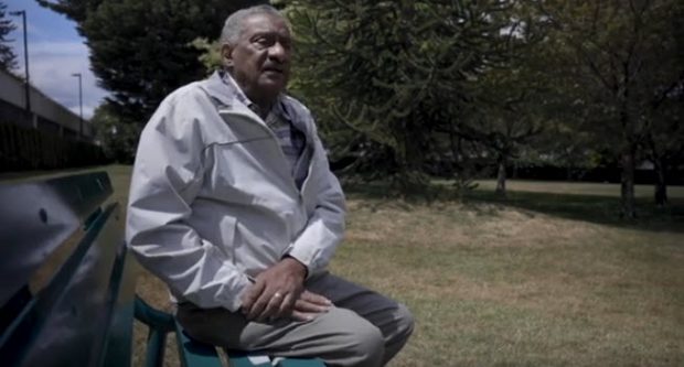 homme âgé, cheveux poivre et sel et moustache, assis sur un banc dans un parc gazonné, tournée vers la caméra. Arbres à feuilles persistantes en arrière-plan