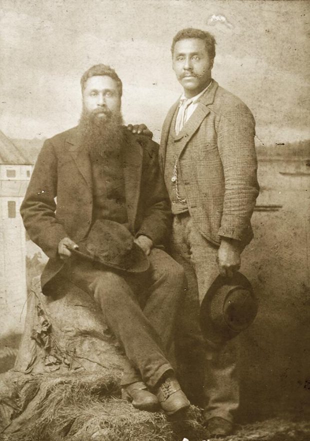 photo couleur sépia, homme assis sur un rocher portant une barbe pleine et tenant un chapeau; un autre homme est debout à ses côtés, rasé et tenant un chapeau; derrière, un paysage marin