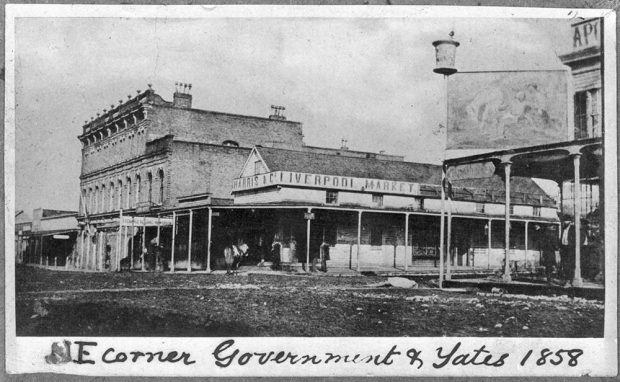 Le texte du bas indique « NE corner Government & Yates 1858 » (angle nord-est, rues Government et Yates, 1858). Intersection de deux rues en terre battue montrant des bâtiments commerciaux construits en bois et en brique de longueurs et hauteurs diverses avec différents types de toitures. On peut y voir quelques personnes et des chevaux