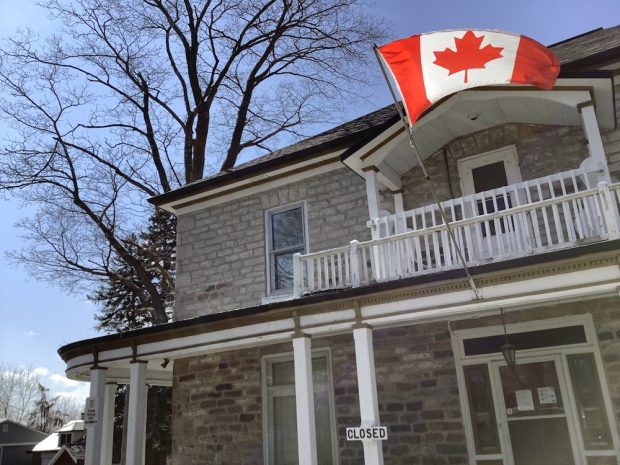 Façade partielle d'un bâtiment en pierre montrant un porche blanc et un balcon au deuxième étage - Un drapeau canadien flotte au sommet de sa porte d'entrée.