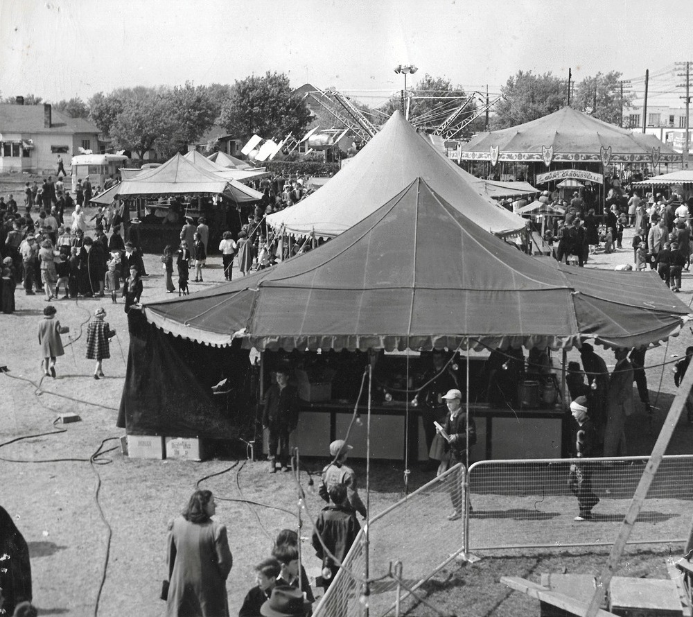 Une photo en noir et blanc d’une foule à une fête foraine parmi de nombreuses tentes; on voit un carrousel en arrière-plan