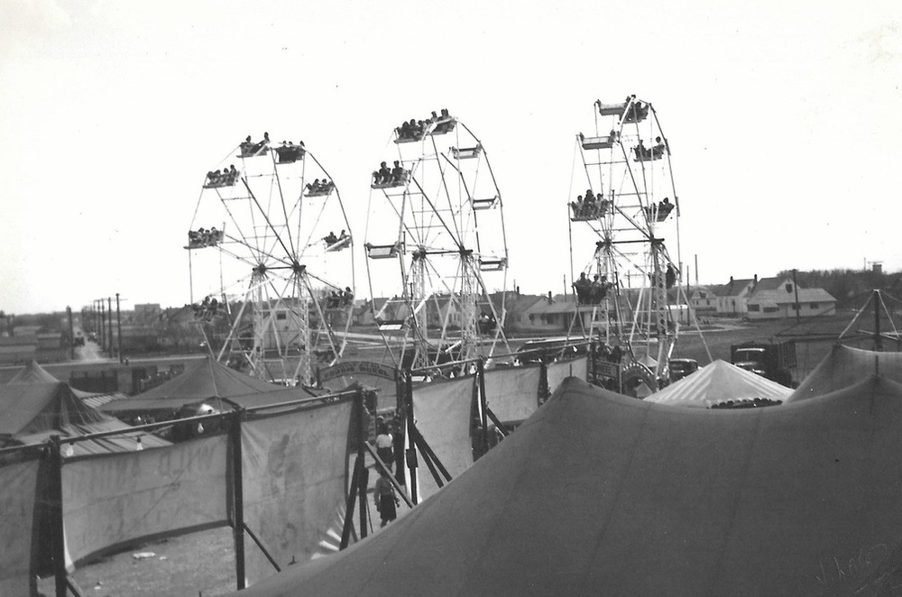 Une photo en noir et blanc d’une fête foraine sur laquelle on voit des gens dans trois grandes roues montées les unes à côté des autres parmi les tentes et les bannières