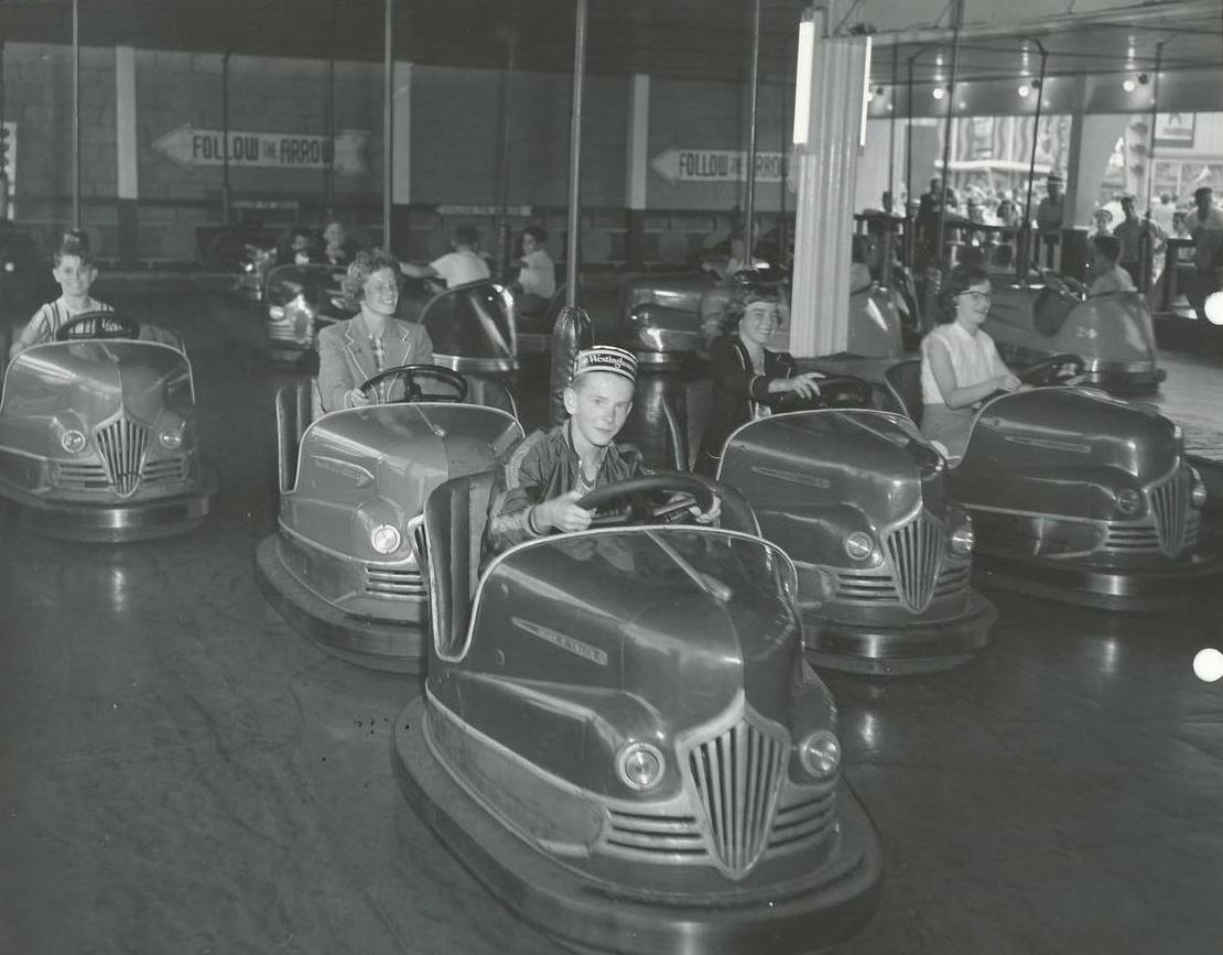 Une photo en noir et blanc de gens qui font un tour d’auto tamponneuse; un jeune garçon conduit une des autos tamponneuses à l’avant