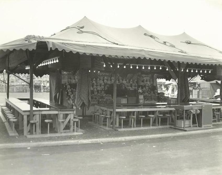 Une photo en noir et blanc d’un stand de bingo installé sous une tente avec des bancs autour des tables, au centre, il y a un mur de peluches et des lumières sont accrochée tout autour du stand