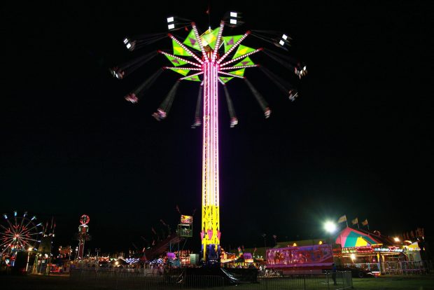 Un manège vertical au centre des attractions, au haut les gens assis sur des chaises attachées par des cordes tournoient; c’est le soir et le manège est illuminé